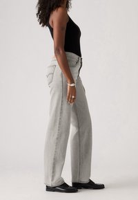 Femme debout de profil portant un jean gris clair taille haute, un top noir sans manches, des chaussures noires et des bagues et un bracelet argentés.