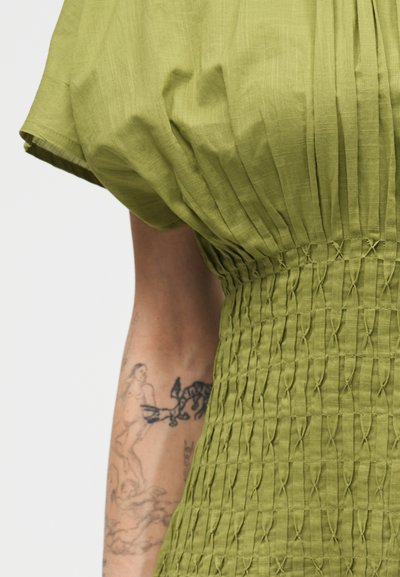 Πράσινο, υφασμάτινο φόρεμα με κοντά μανίκια και σφηκοφωλιά στη μέση, που φοριέται στο αριστερό χέρι και δείχνει τατουάζ μαύρης γραμμής με ανθρώπινες φιγούρες και αφηρημένα σχήματα.