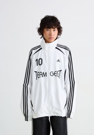 Veste de sport blanche avec des rayures noires et un graphique 'TEAM GEIST', fermeture éclair à l'avant, col haut et détail de logo. Design léger et sportif.