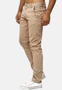 Chino in cotone beige con vestibilità slim, frontale piatto, passanti per cintura e tasche laterali, abbinati a sneaker bianche con dettagli rossi.