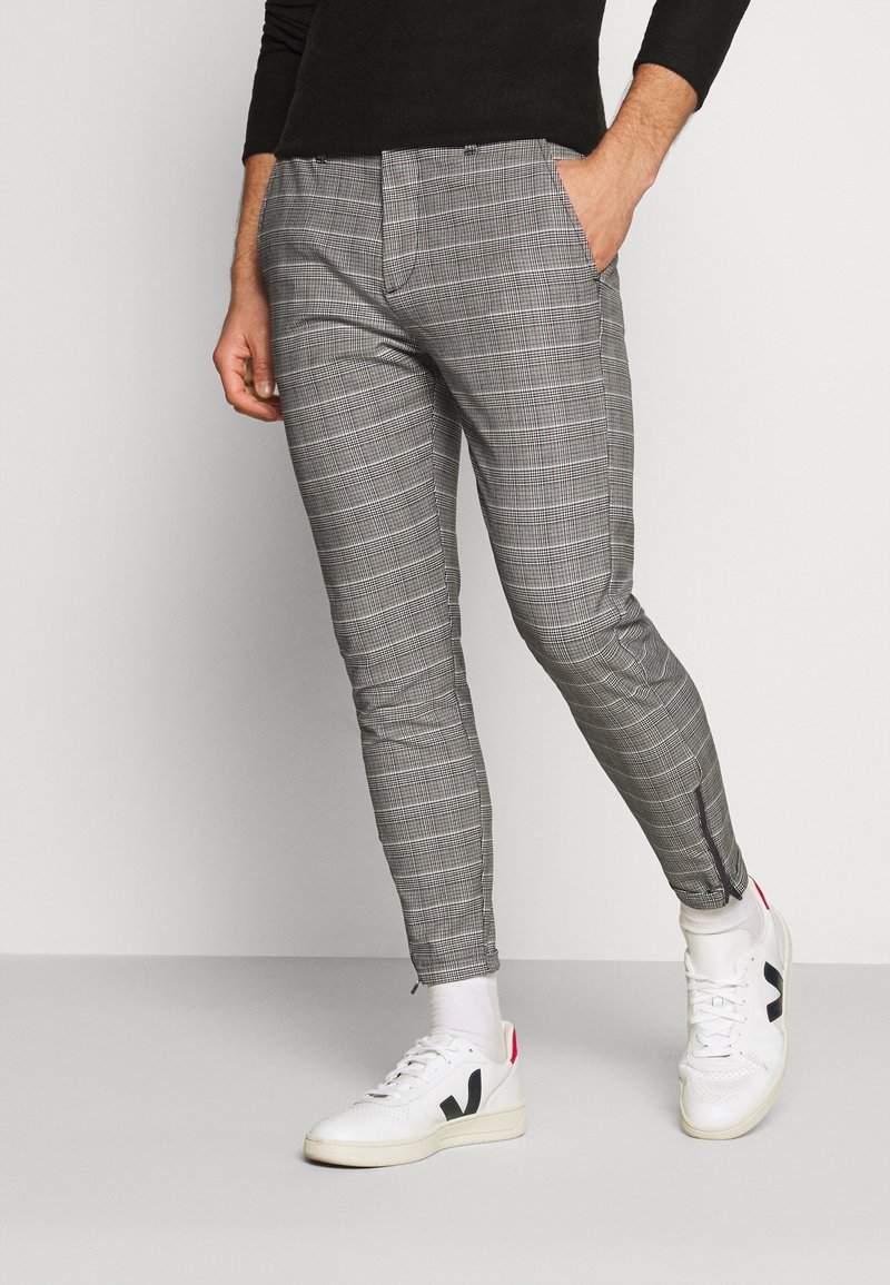 Homme portant un pantalon slim à carreaux gris avec des zips aux chevilles, des chaussettes blanches, des baskets blanches et un t-shirt noir à manches longues, debout devant un fond uni.