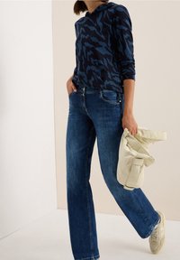 Hoodie marine avec motif abstrait bleu, jean en denim et baskets beige. Sac crème tenu dans la main gauche. Tenue décontractée avec des textures superposées.