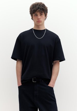 Fiatal férfi göndör hajjal, fekete túlméretezett pólót, fekete nadrágot és ezüst láncnyakláncot visel, kezei a zsebében, fehér háttér előtt áll.