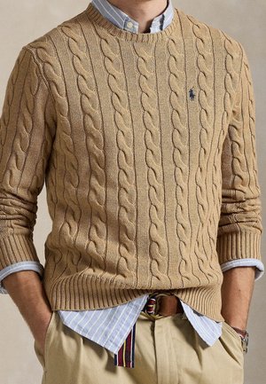 Homme portant un pull beige en maille torsadée sur une chemise rayée bleue, un pantalon kaki et une ceinture rayée, les mains dans les poches.