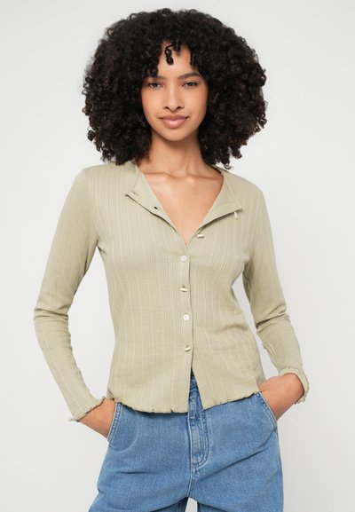 Svetlo zelena rebrasta dolga srajca z gumbi in ovratnikom z raztrganim robom, v kombinaciji z modrimi kavbojkami visoko pasu.