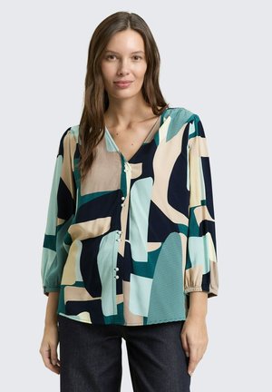 Femme portant un chemisier à manches longues boutonné avec des motifs abstraits beige, vert sarcelle, noir et crème, assorti à un pantalon foncé.