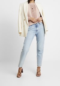 Person, die eine hellpinkfarbene Bluse, ein cremefarbenes Blazer über die Schultern drapiert, hellblaue Jeans, braune Riemchen-Heels und mehrschichtige Halsketten trägt.