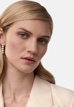 Lyse beige blazer over en lys hudtone. Har langt, glat blondt hår og hængende perleøreringe. Neutral makeup med definerede øjne.