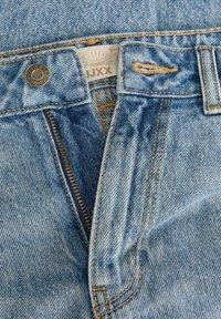 Sinine teksapükside lähivõte, mille tõmblukk on osaliselt avatud, metallist nööp tähtedega ja vöökoha sees nähtav kangasesilt.