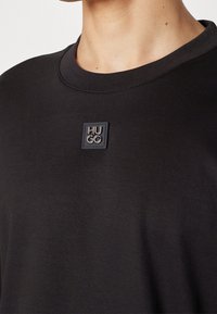 Zwarte t-shirt met ronde halslijn, met een vierkante, geperste logopatch met de tekst "HUGO" op de borst. Gladde stofstructuur.