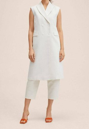 Frau trägt ärmellose weiße, doppelreihige lange Weste, weiße kurze Hose und orangefarbene Sandalen mit Absatz, steht vor einfarbigem beigem Hintergrund.