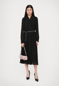 Γυναίκα που φοράει ένα μαύρο πλισέ φόρεμα με μακριά μανίκια και αλυσίδα στη μέση, κρατώντας μια μικρή ροζ τσάντα και μαύρες ψηλές γόβες με μυτερή μύτη, σε άσπρο φόντο.