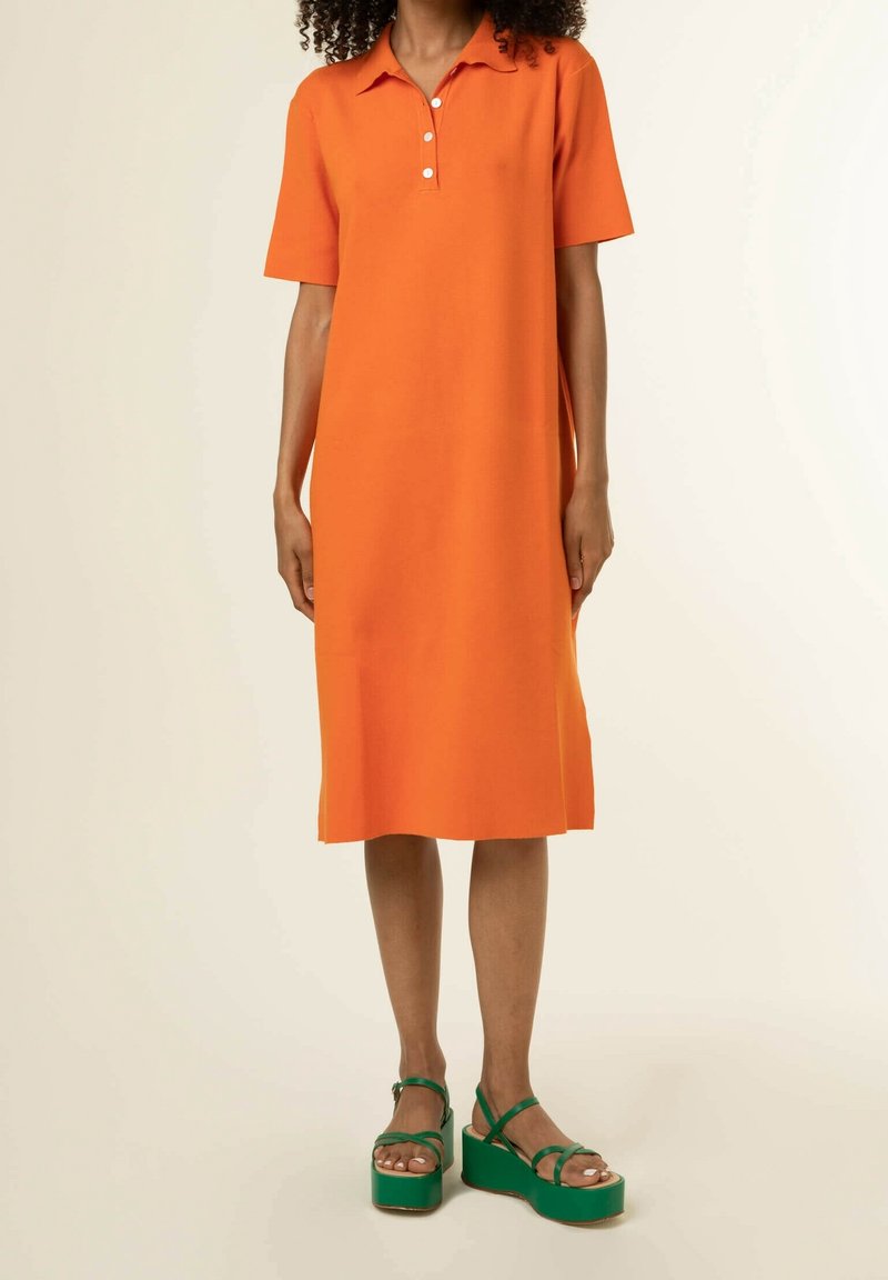 Femme portant une robe orange mi-longue à manches courtes et des sandales à plateforme vertes avec des brides à la cheville.