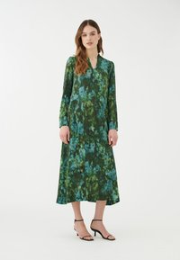 Abito floreale al ginocchio in tonalità di verde e blu, realizzato in tessuto leggero con maniche lunghe e scollatura a V. Sandali neri con cinturini.