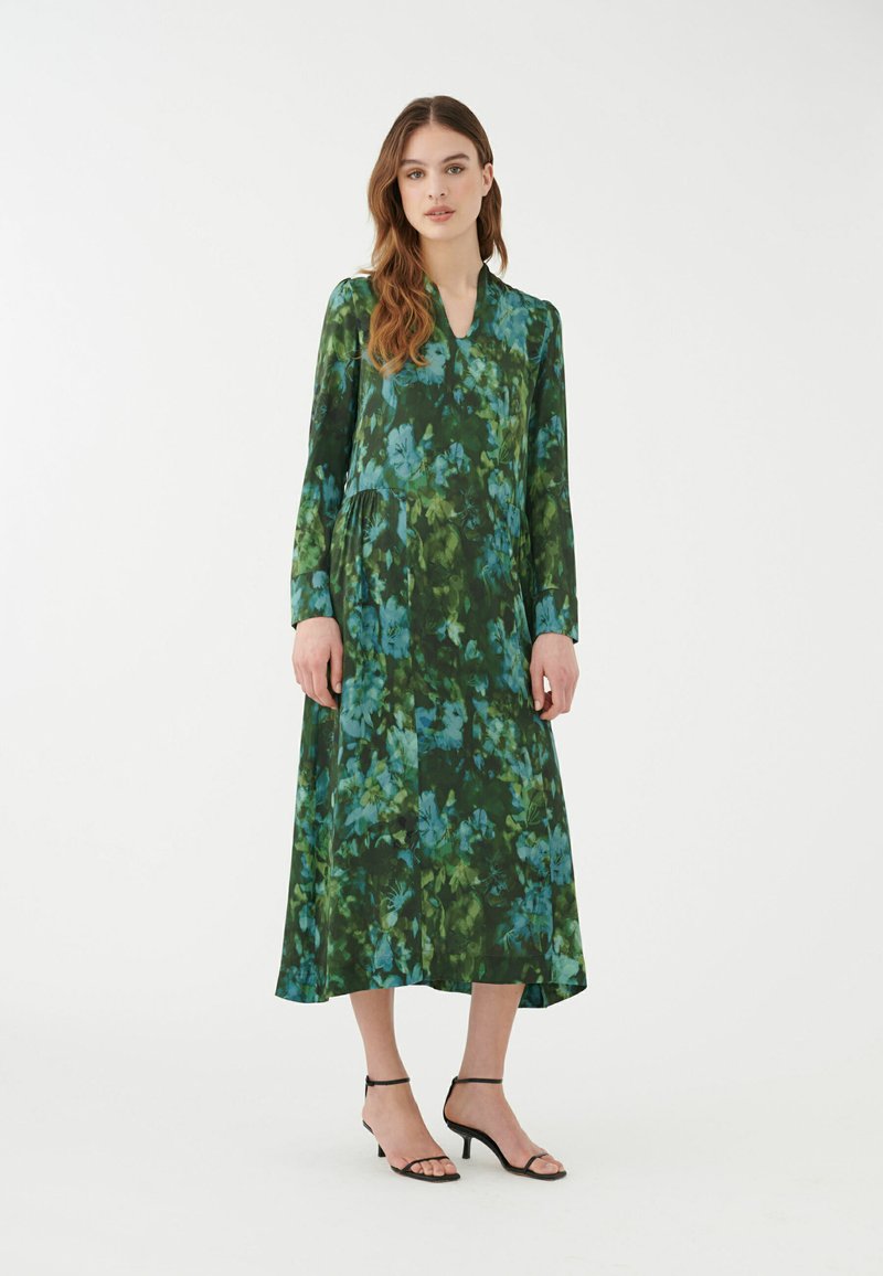 Abito floreale al ginocchio in tonalità di verde e blu, realizzato in tessuto leggero con maniche lunghe e scollatura a V. Sandali neri con cinturini.