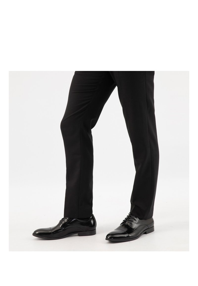 Beine in schwarzen Anzughosen und polierten schwarzen Leder-Herrenschuhen, die auf einer weißen Oberfläche stehen.