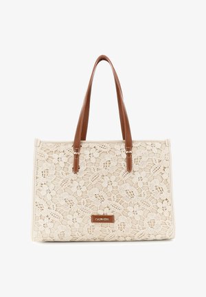 Beige kangaskassi, jossa on kukallinen pitsipäällys, ruskeat nahkakahvat ja pieni ruskea brändin nimilappu keskellä etupuolella.