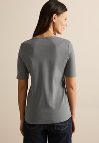 T-shirt grigio a maniche corte con scollatura tonda, texture liscia e orlo leggermente curvo, abbinato a jeans in denim scuro.