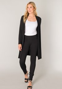 Zwarte lange cardigan over een wit hemdje, gecombineerd met zwarte legging en zwarte sandalen met hak. De cardigan heeft een open ontwerp en een gladde textuur.