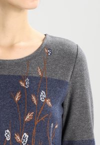 Grå tröja med marinblå rand, prydd med blommig broderi i vitt och brunt. Mjuk textur, rund halsringning och långa ärmar.