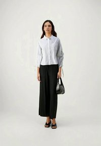 Witte overhem shirt met opgerolde lange mouwen, gecombineerd met zwarte geplooide wijde broek en zwarte open teen sandalen, met een zwarte tas.