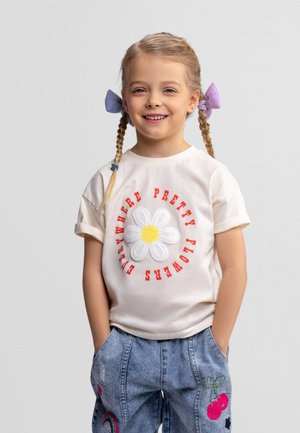 Uśmiechnięta dziewczynka z warkoczami związanymi fioletowymi kokardami, ubrana w białą koszulę z stokrotką i czerwonym napisem oraz niebieskie dżinsy z kolorowymi naszywkami.