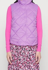 Gilet matelassé violet avec col haut et fermetures à pression, superposé sur un haut à manches longues rose et une jupe à motif floral.