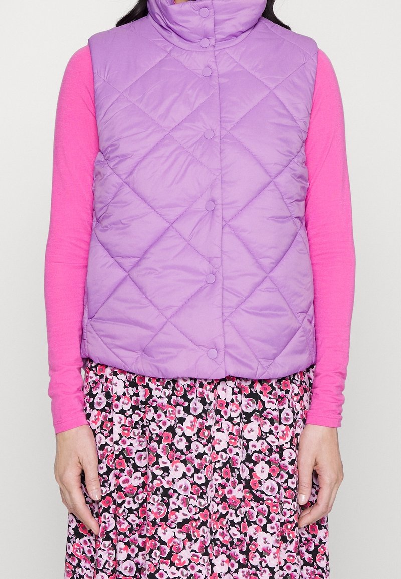 Gilet matelassé violet avec col haut et fermetures à pression, superposé sur un haut à manches longues rose et une jupe à motif floral.