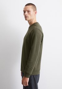 Olíva zöld, kerek nyakú pulóver puha, texturált anyagból. Bordás mandzsettákkal és alsó szélekkel rendelkezik, minimális márkajelzéssel a mellen.