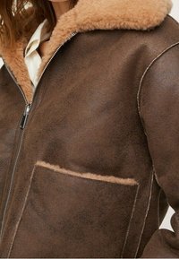 Veste en shearling marron avec un grand col doux, une fermeture éclair à l'avant et une poche visible, portée sur une chemise blanche.