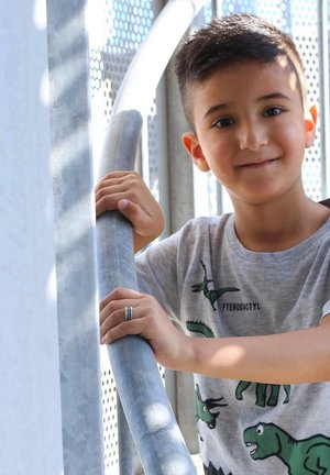 Kind in een grijs T-shirt met groene dinosaurussen, die een zilveren metalen leuning vasthoudt; gestructureerde achtergrond met licht dat erdoorheen filtert.