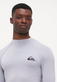 Világosszürke, hosszú ujjú rash guard rugalmas anyagból, fekete logóval a bal felső részen és laposvarrásos részletekkel.