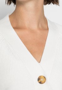 Cardigan bianco lavorato a maglia con scollatura a V e un grande bottone rotondo in ambra. Il tessuto presenta un motivo texture con dettagli sottili.