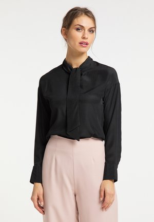 Vrouw die een zwarte blouse met lange mouwen en een halsstrik draagt, gecombineerd met lichtbeige, hooggetailleerde broek, staand tegen een effen witte achtergrond.