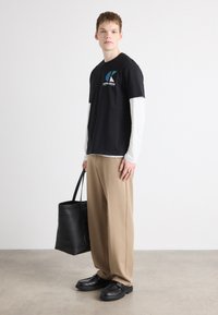 Sort grafisk T-shirt over en hvid langærmet skjorte, beige bukser og sorte sko. Holder en sort tote bag. Enkel, moderne design.