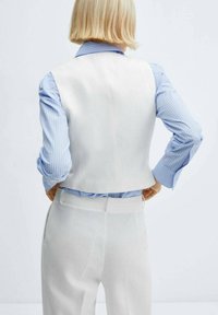 Gilet bianco su misura con design cropped, indossato sopra una camicia a righe blu chiaro, abbinato a pantaloni leggeri dello stesso colore.