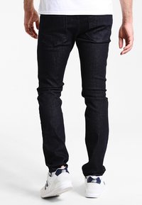 Jeans de mezclilla de lavado oscuro con corte slim, que cuentan con dos bolsillos traseros y costuras sutiles. Llevados con zapatillas deportivas blancas.