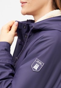 Mörkblå vattentät jacka med krage klädd i fuskpäls, med en logotyp på ärmen och dragsnören vid halsen för justering.