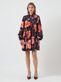 Czarna sukienka w kwiaty z dużymi pomarańczowymi kwiatami, długimi bufkami, kołnierzykiem zapinanym na guziki oraz warstwowym krojem. Zestawiona z czarnymi szpilkami.