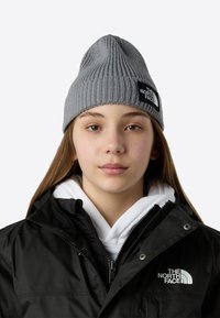 Γκρι πλεκτός σκούφος με σχέδιο κορυφής και ριμπ ύφασμα, με μαύρη ορθογώνια ετικέτα με λευκό κείμενο. Συνδυασμένος με μαύρο μπουφάν και λευκό φούτερ με κουκούλα.