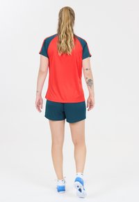 Kvinde der står med ryggen til kameraet iført rød og mørk teal sportstøj, hvide og blå sneakers, med synlige arm tatoveringer og en hestehale.