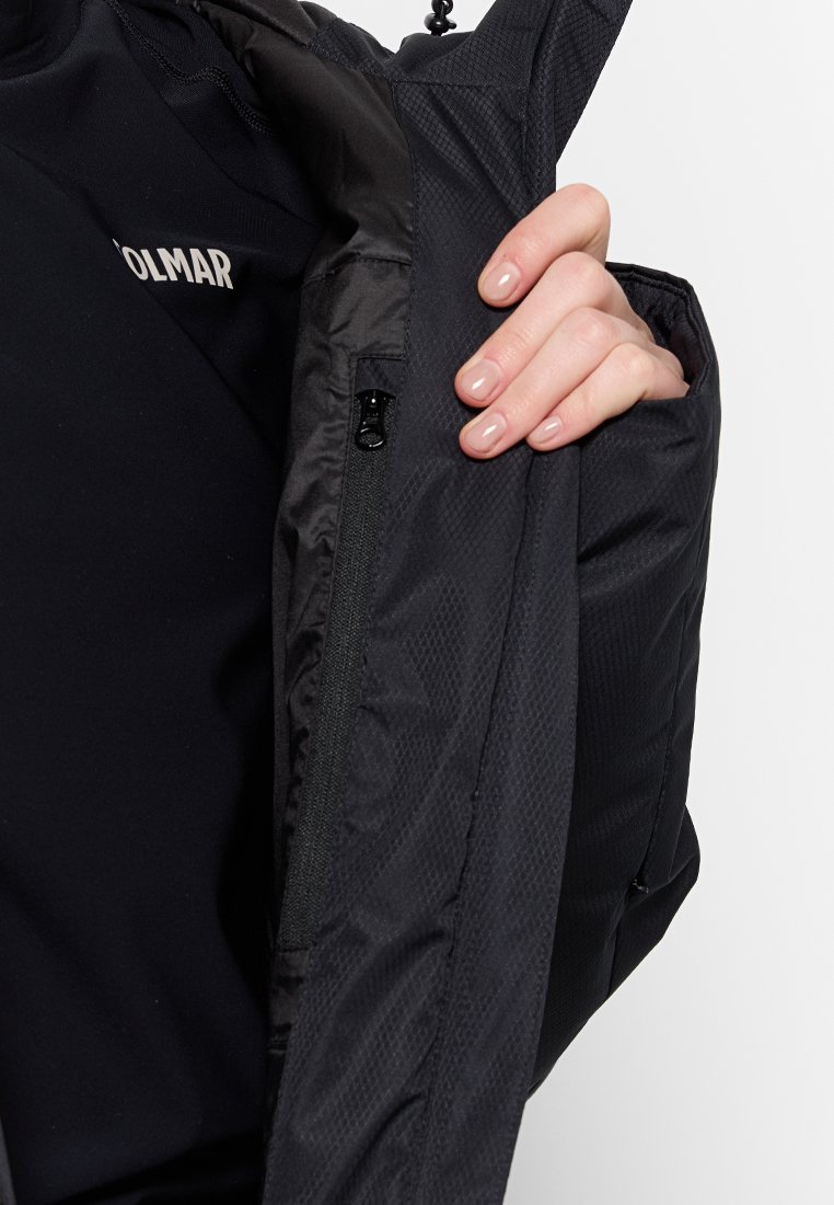 Mano che tiene aperta una giacca nera mostrando la tasca interna con zip e la texture del tessuto, persona che indossa una maglietta nera con lettere bianche parziali.