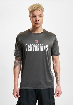 Tummanharmaa urheilupaita, jossa on pyöreä kaula-aukko ja lyhyet hihat. Paidassa on valkoisella tekstillä "COLOGNE CENTURIONS" ja punainen kypärälogo.