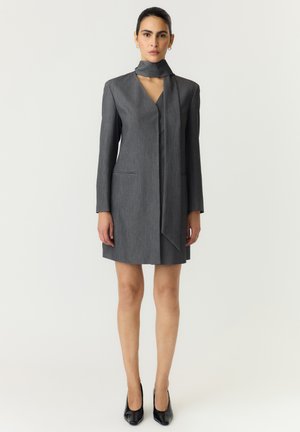 Femme debout portant une robe grise à manches longues avec un détail foulard au cou et des chaussures à talons noires sur un fond uni.