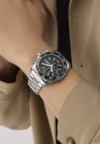 Zegarek chronograf ze stali nierdzewnej z czarną tarczą, srebrnymi akcentami i bransoletą, z trzema sub-tarczami i fluorescencyjnymi znacznikami.