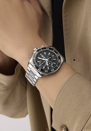 Nerūsējošā tērauda hronogrāfu pulkstenis ar melnu ciparnīcu, sudraba akcentiem un siksnu ar rokassprādzi, kas ietver trīs subciparnīcas un gaismu atstarotājus.