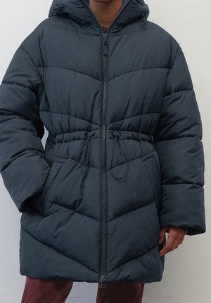 Person wearing a black quilted puffer jacket with a front zipper, drawstring waist, and side pockets, standing against a plain wall.