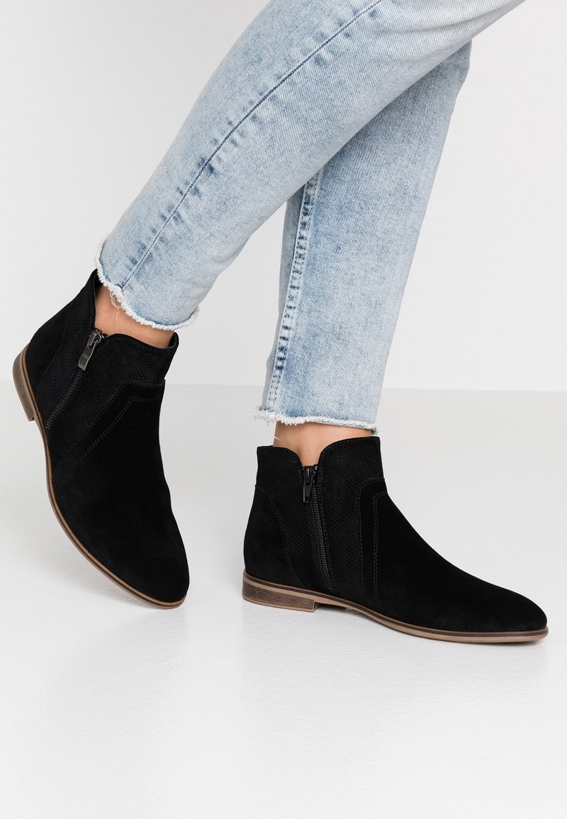 Personne portant un jean bleu clair à ourlet effiloché et des bottines noires en daim avec des fermetures éclair latérales sur un sol blanc.