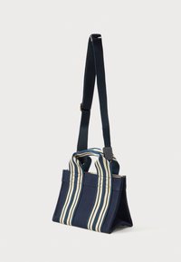 Ναυτικό μπλε τσάντα ώμου με κατακόρυφες λευκές και ναυτικές ρίγες στις λαβές και ρυθμιζόμενο ναυτικό ιμάντα, σε όρθια θέση σε απλό φόντο.