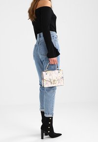 Blomsterprydet håndtaske med rektangulær form, beige bund og metalliske accenter; parret med høj talje blå jeans og sorte hælsko.